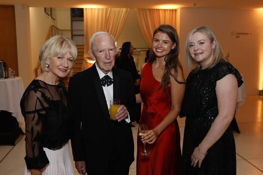 Four people pose together at a reception, dressed formally for the UCD Alumni Awards; Peter Timoney in a tuxedo holds a glass of orange juice beside guests in black and red gowns.